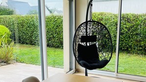 Comedor con mesa de madera, sillas, una mecedora y vistas a un jardín a través de grandes ventanales.