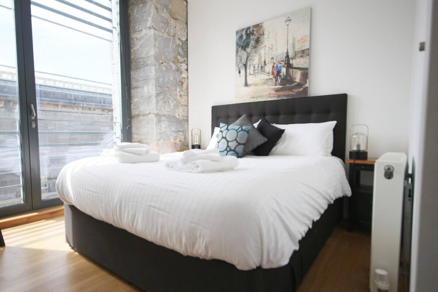 A modern bedroom with a large bed, a painting on the wall, and a view through the window.