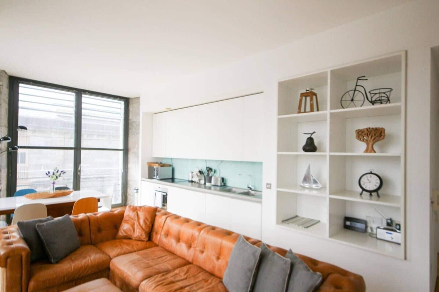 A modern living room with a leather sofa, a dining area with a table and chairs, and a white shelving unit with decorative items.