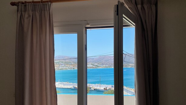 A room with a sliding glass door leading to a balcony overlooking a coastal area with buildings and a mountain in the distance. - Creta Sun Apartments (Ierapetra)