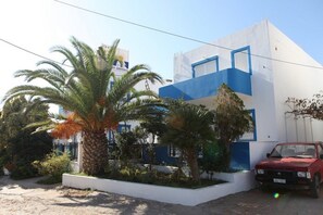 A building with a white and blue facade, palm trees, and a red car parked in front. - Creta Sun Apartments (Ierapetra)