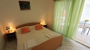 Un dormitorio con cama, dos almohadas, una mesita de noche con lámpara, un cuadro en la pared y una ventana con cortinas traslúcidas.