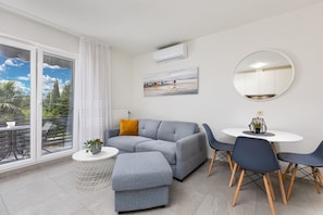 Uma sala de estar moderna com um sofá seccional cinza, uma mesa redonda pequena com cadeiras, uma janela grande com vista para vegetação e um quadro na parede.
