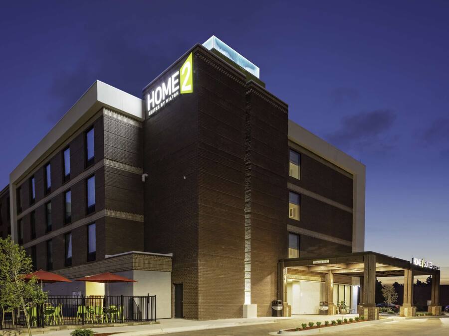 Ett modernt hotell med en tydlig skylt som visar "HOME 2", ett takförsett utomhusområde med sittplatser och en väl upplyst entré.