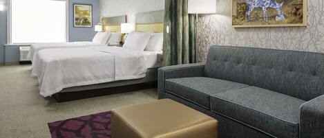 A hotel room with a bed, a grey sofa, a small ottoman, and a painting of a blue deer on the wall.