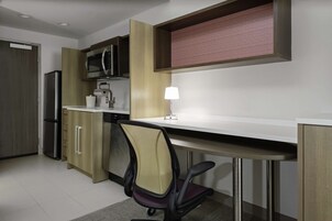 A modern kitchen with a built-in microwave, a refrigerator, and a coffee maker. There is a desk with a chair and a lamp.