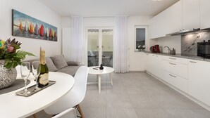 A modern kitchen with white cabinets, a black microwave, and a stainless steel range hood. There is a dining area with a white table and chairs, and a painting of sailboats on the wall.