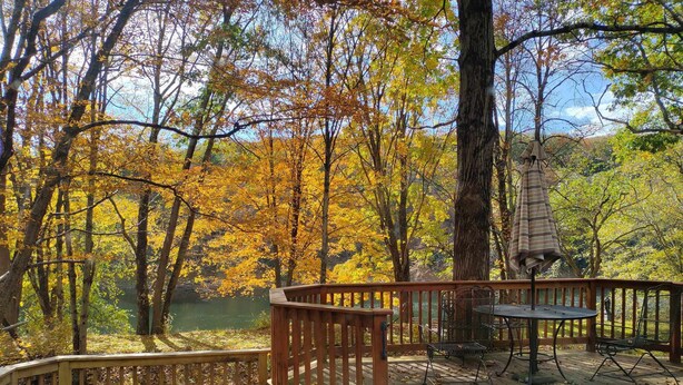 Una terraza de madera con una mesa y sillas, con vista a un lago rodeado de árboles de colores otoñales.