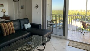 A living room with a black leather sofa, a glass coffee table, and a sliding glass door leading to a balcony with a view of greenery. - Beach, Shelling, Water Sport, Restaurant Right Across The Street!  Paradise!! (Bonita Springs)