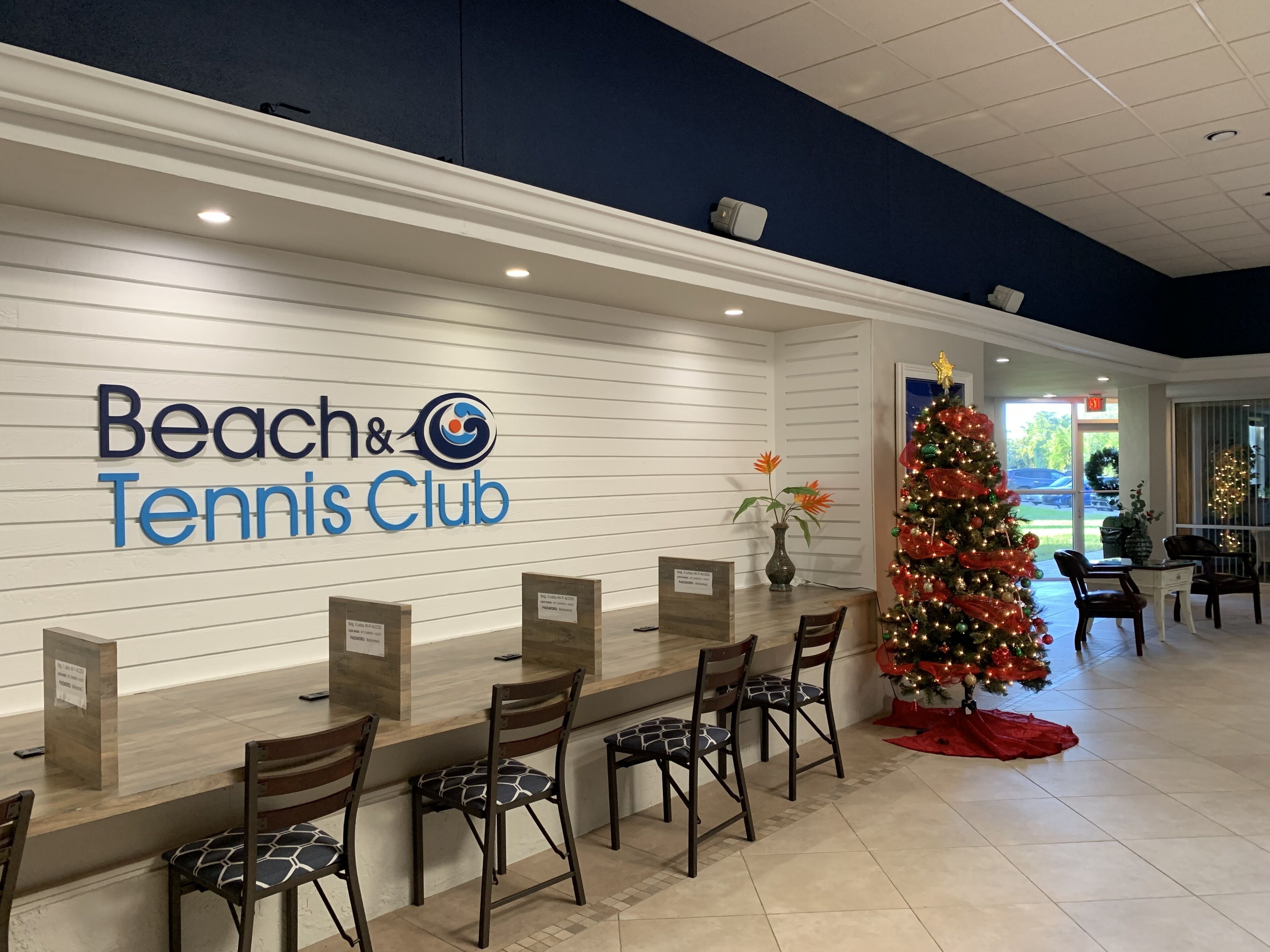 Un espace d’accueil avec un panneau indiquant « Club de plage et de tennis », un sapin de Noël et un coin salon avec des chaises et des tables.