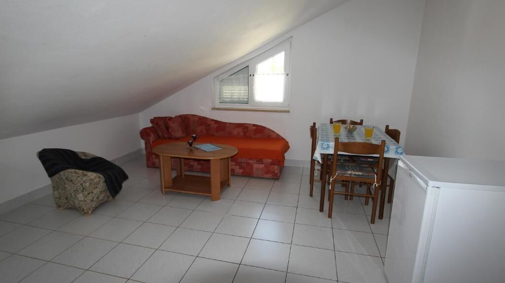 Une petite pièce bien éclairée, avec un sol carrelé, un canapé rouge, une table en bois avec des chaises, une petite table avec une lampe et un réfrigérateur blanc.