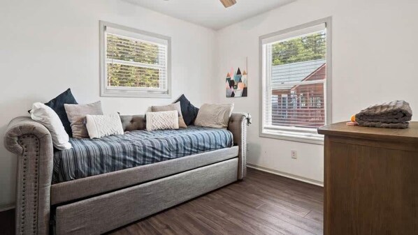 Une pièce comprenant un canapé-lit, deux fenêtres équipées de stores, une petite table et un ventilateur de plafond.