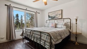 A bedroom with a bed, a window with curtains, a ceiling fan, a bedside lamp, and a painting on the wall. - Imogene Home — 4BR + Hot Tub, Sleeps 10, 15 min to Ski Cooper, Ping Pong Garage (Leadville)