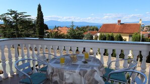 A balcony with a table set for two, overlooking a coastal town with red-roofed buildings and mountains in the distance.