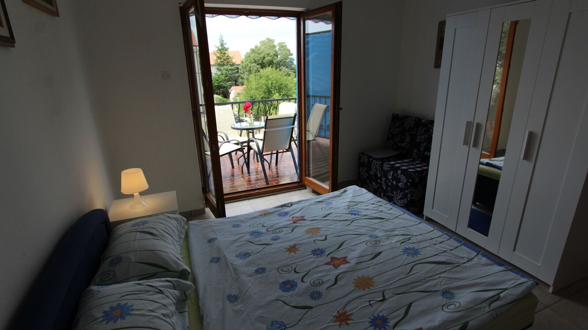 Un dormitorio con cama, mesita de noche, lámpara, armario y una puerta corrediza de vidrio que da a un patio exterior con mesa y sillas.