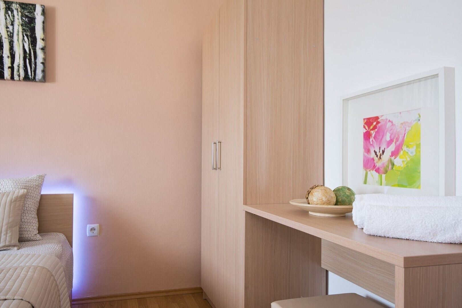 Un dormitorio moderno con un armario de madera, una mesita de noche con una bandeja de fruta y una toalla, y un cuadro colgado en la pared.