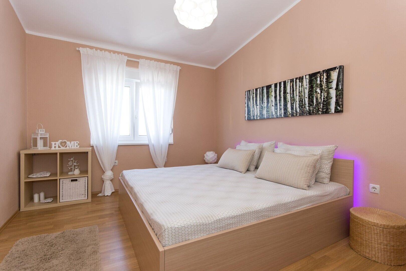Ein Schlafzimmer mit einem Bett, einem Nachttisch, einer Lampe, einem Fenster mit Vorhängen, einem Wandbild und einem kleinen Korbstuhl.