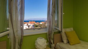 A room with a bed, a window with curtains, and a view of buildings and the sea. - Apartment Jana in Malinska (Malinska)