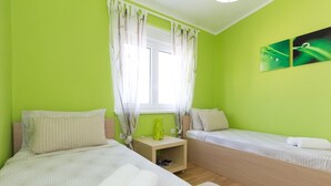 Ein Zimmer mit zwei Betten, einem Fenster mit durchsichtigen Vorhängen, einem kleinen Tisch und einer grünen Wand.
