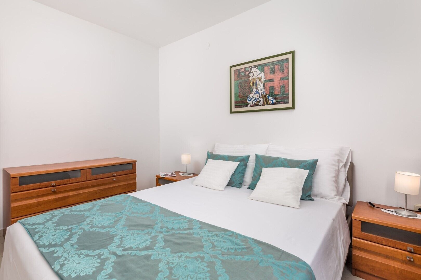 A bedroom with a bed, two bedside tables, a wooden dresser, and a framed artwork on the wall.