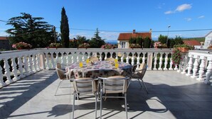 Mesa redonda con sillas en una terraza con barandilla blanca, con vista a una zona residencial con casas y árboles.