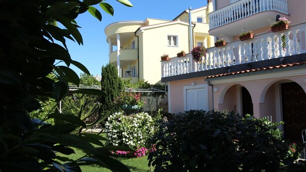 Un edifici de diverses plantes amb balcons, una zona de jardí amb diverses plantes i un cel blau clar.