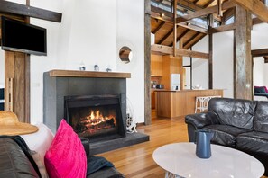 A living room with a fireplace, a black leather sofa, a white coffee table, and a kitchen area in the background.