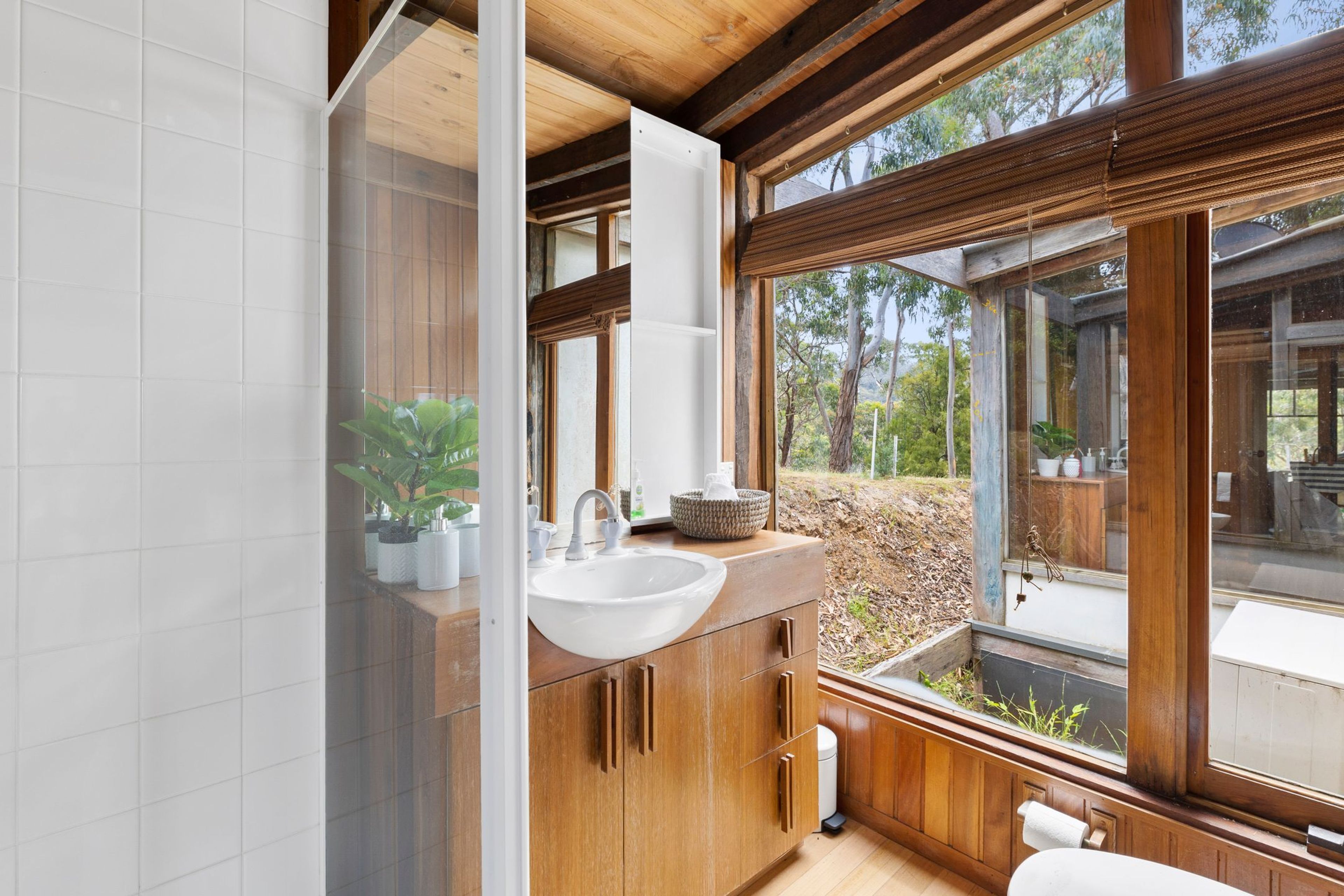 A modern bathroom with a wooden vanity, a white sink, and a large window offering a view of the outdoors.