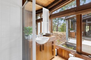 Une salle de bain moderne avec un meuble-lavabo en bois, un lavabo blanc et une grande fenêtre donnant sur l’extérieur.