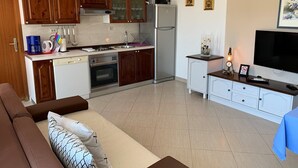 Een praktische keuken met houten kastjes, een koelkast, een magnetron en een fornuis. Er is een eethoek met een tafel en stoelen, en een woonkamer met een bank en een televisie.