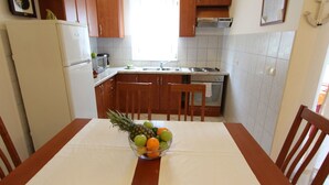 Eine Küche mit Holzausstattung, einem weißen Kühlschrank, einer Mikrowelle und einem Spülbecken. In der Mitte steht ein Tisch mit einer Schüssel Obst.