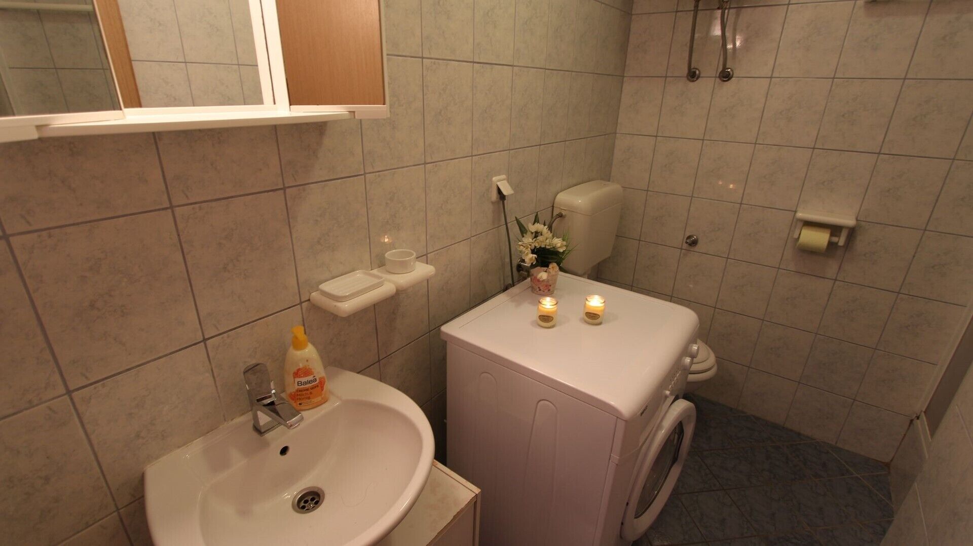 Un baño con lavamanos, inodoro y lavadora. Hay velas sobre el inodoro, una botella de loción y una planta pequeña.
