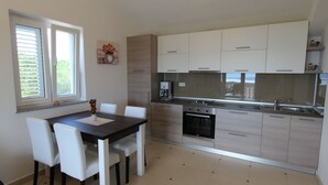 A modern kitchen with white cabinets, a built-in oven, and a coffee maker. There is a dining table with chairs and a window with blinds. - Deluxe apartment Jela 2 for 4 people with sea view (Sveti Vid Miholjice)