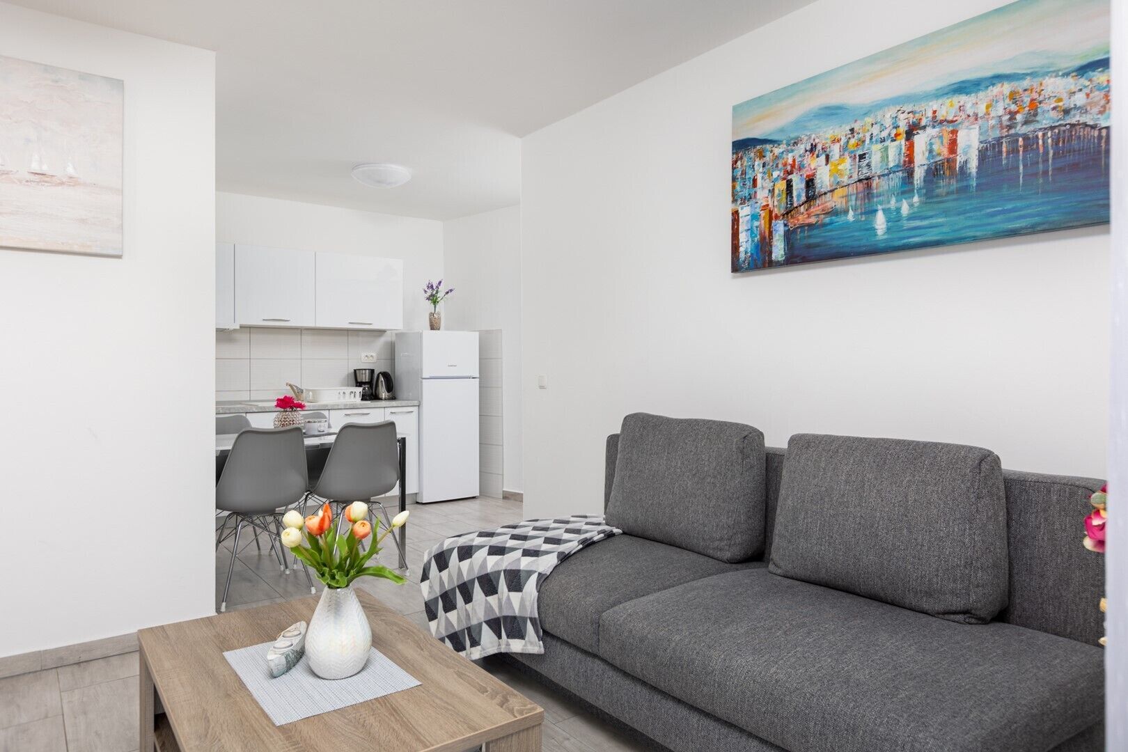A modern living room with a grey sofa, a wooden coffee table with a vase of flowers, and a painting of a cityscape on the wall.