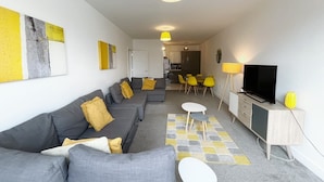 A modern living room with a grey sofa, yellow cushions, a TV, and a dining area with yellow chairs. - Anstey Heights - Apartment 8 - Hopewell (Easton)