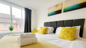 A hotel room with a large bed, a window with curtains, a potted plant, and two wall art pieces. - Anstey Heights - Apartment 8 - Hopewell (Easton)