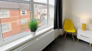 A room with a large window, a radiator, a yellow chair, and a white bedside table with a lamp. - Anstey Heights - Apartment 8 - Hopewell (Easton)