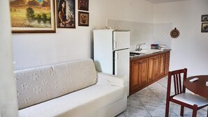 A compact living space with a sofa, a small kitchen area with a refrigerator and sink, wooden cabinets, and a dining table with a chair. - Appartamento Iris 2- IUN P2186 Rose (Cala Gonone)