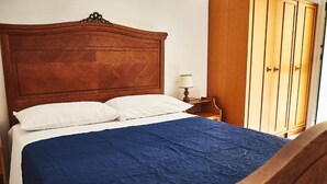 A bed with a wooden headboard, a blue bedspread, and white pillows. There is a wooden nightstand with a lamp to the right of the bed. A wooden wardrobe is visible in the background. - Appartamento Iris 2- IUN P2186 Rose (Cala Gonone)