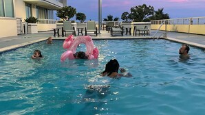 Ein Schwimmbad mit Personen, einem rosa Schwimmreifen und einem Gebäude mit Balkonen im Hintergrund.