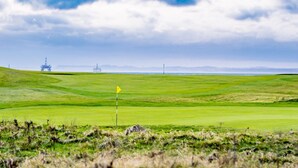 Un campo de golf con un mástil de bandera, plataformas petrolíferas a lo lejos y un cielo despejado.