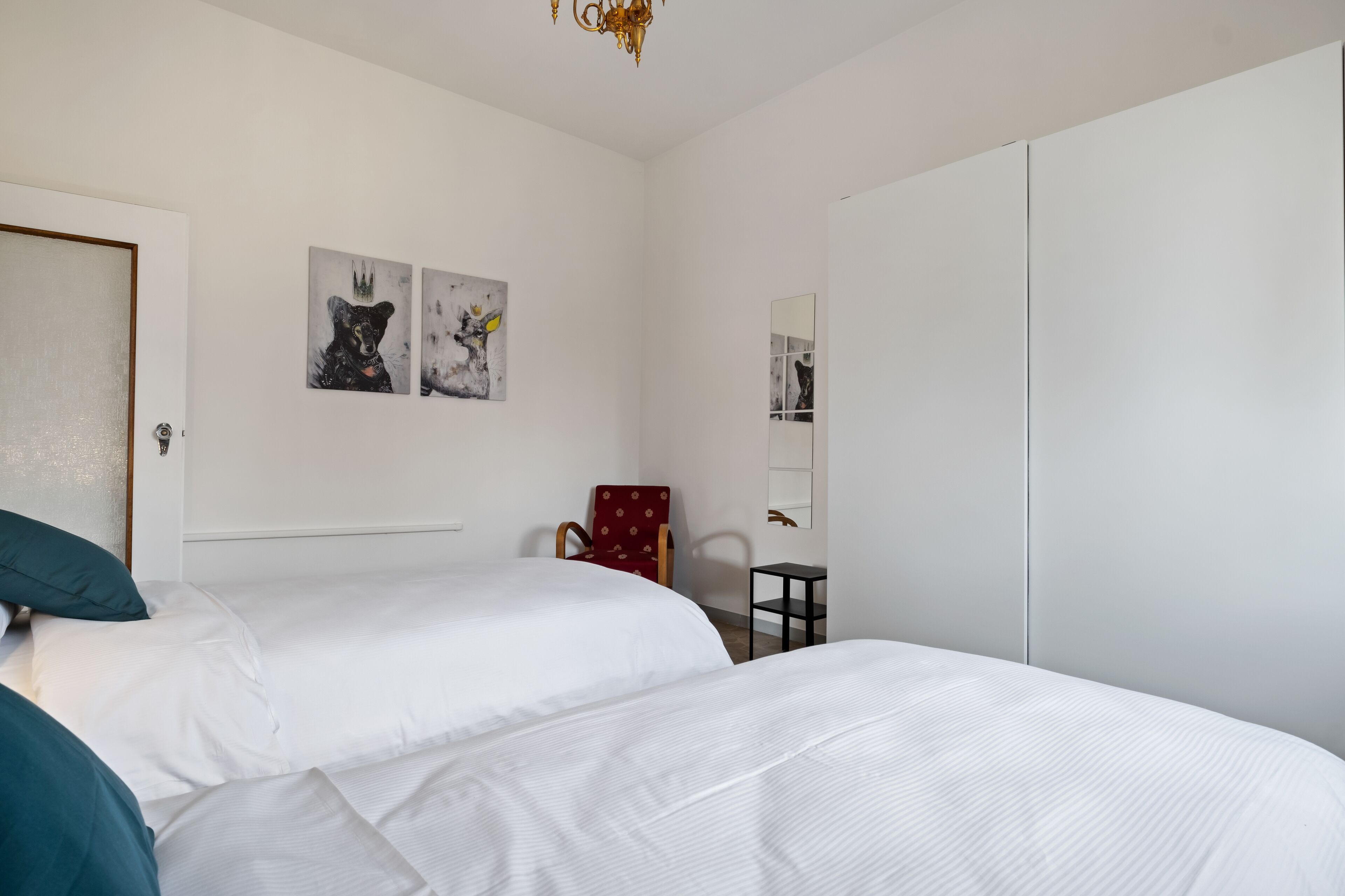A bedroom with two beds, a chair, a small table, and artwork on the wall.