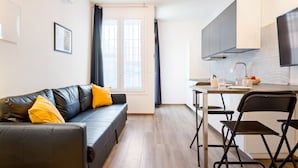 Un apartamento moderno con sofá de cuero negro, una mesa de comedor con sillas, zona de cocina y un gran ventanal con cortinas.