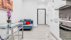 A modern apartment with a glass dining table, a blue sofa with red cushions, a kitchen with white cabinets, and a cityscape painting on the wall.