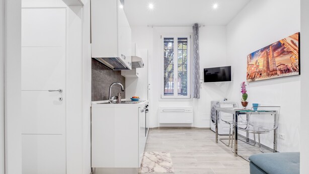 A compact kitchen with a built-in stove, a window with curtains, a TV mounted on the wall, and a dining area with a table and chairs.