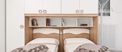 Une chambre avec deux lits, une tête de lit en bois, une étagère intégrée remplie de livres et un radiateur.
