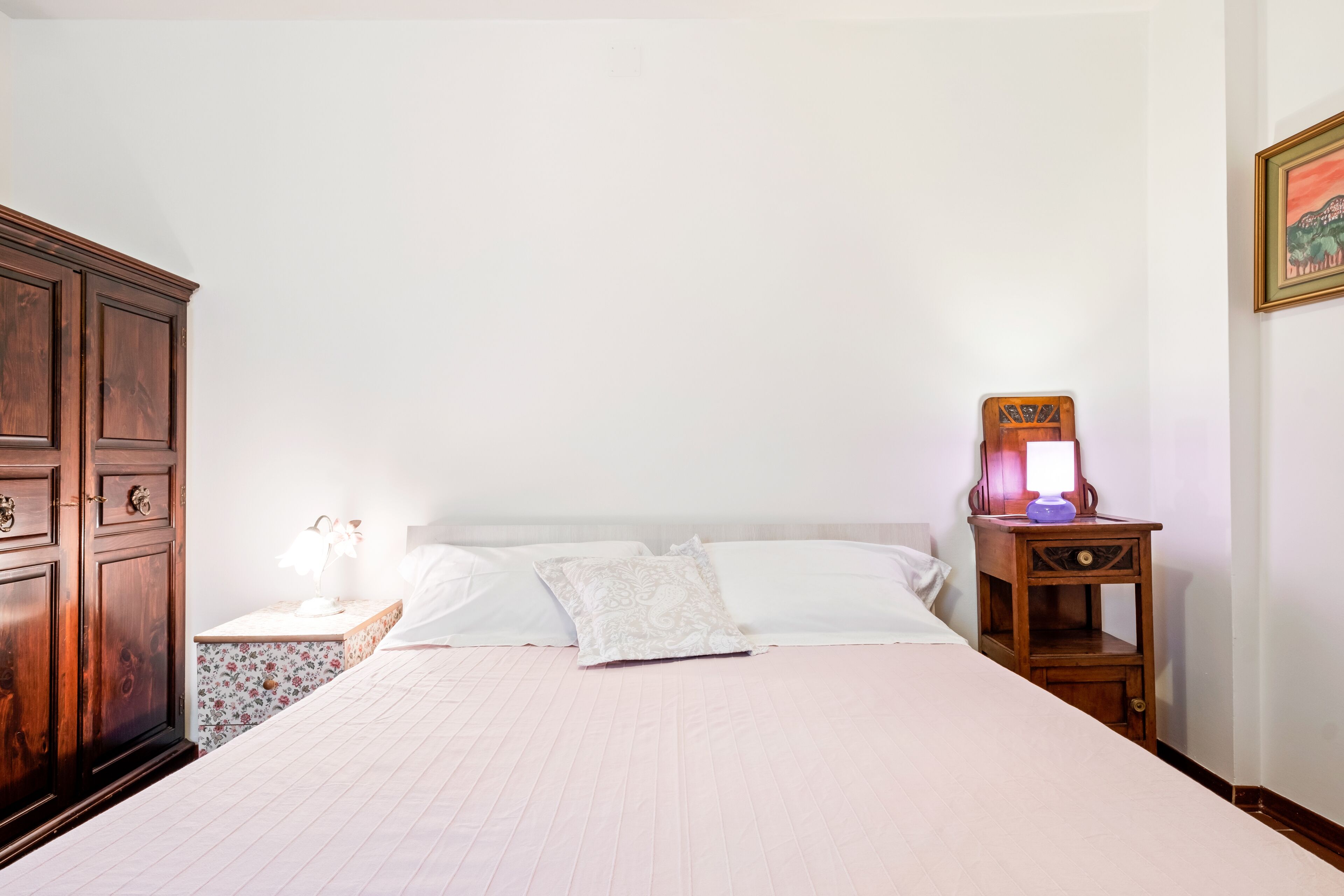 A neatly made bed with white linens, a wooden nightstand with a lamp, and a wooden wardrobe.