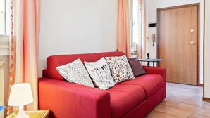 Ein rotes Sofa mit gemusterten Kissen, ein kleiner Tisch mit einer Lampe und eine Holztür in einem gut beleuchteten Raum mit Vorhängen.