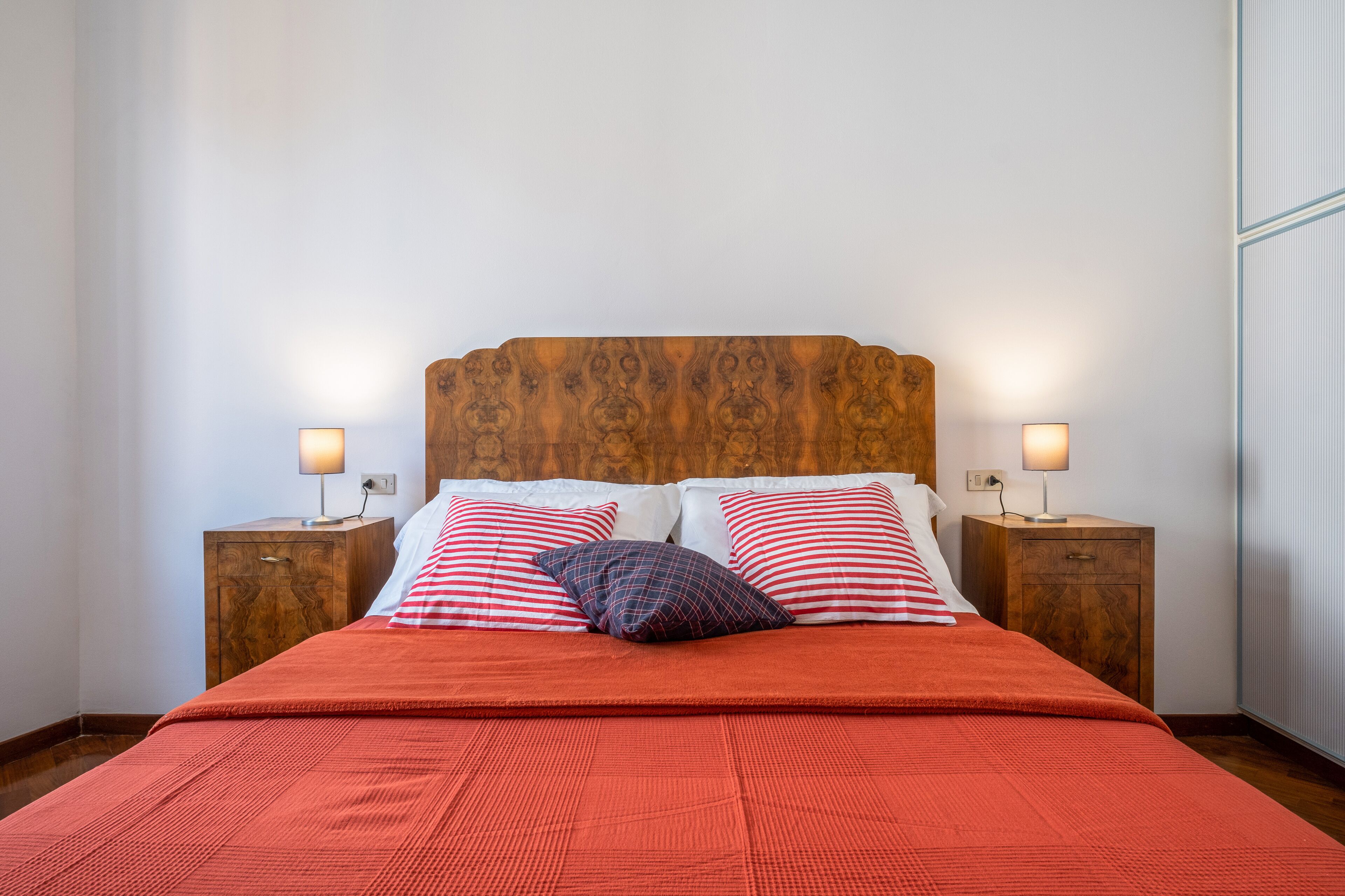 A neatly made bed with a red blanket, two pillows with red and white stripes, and two bedside tables with lamps.