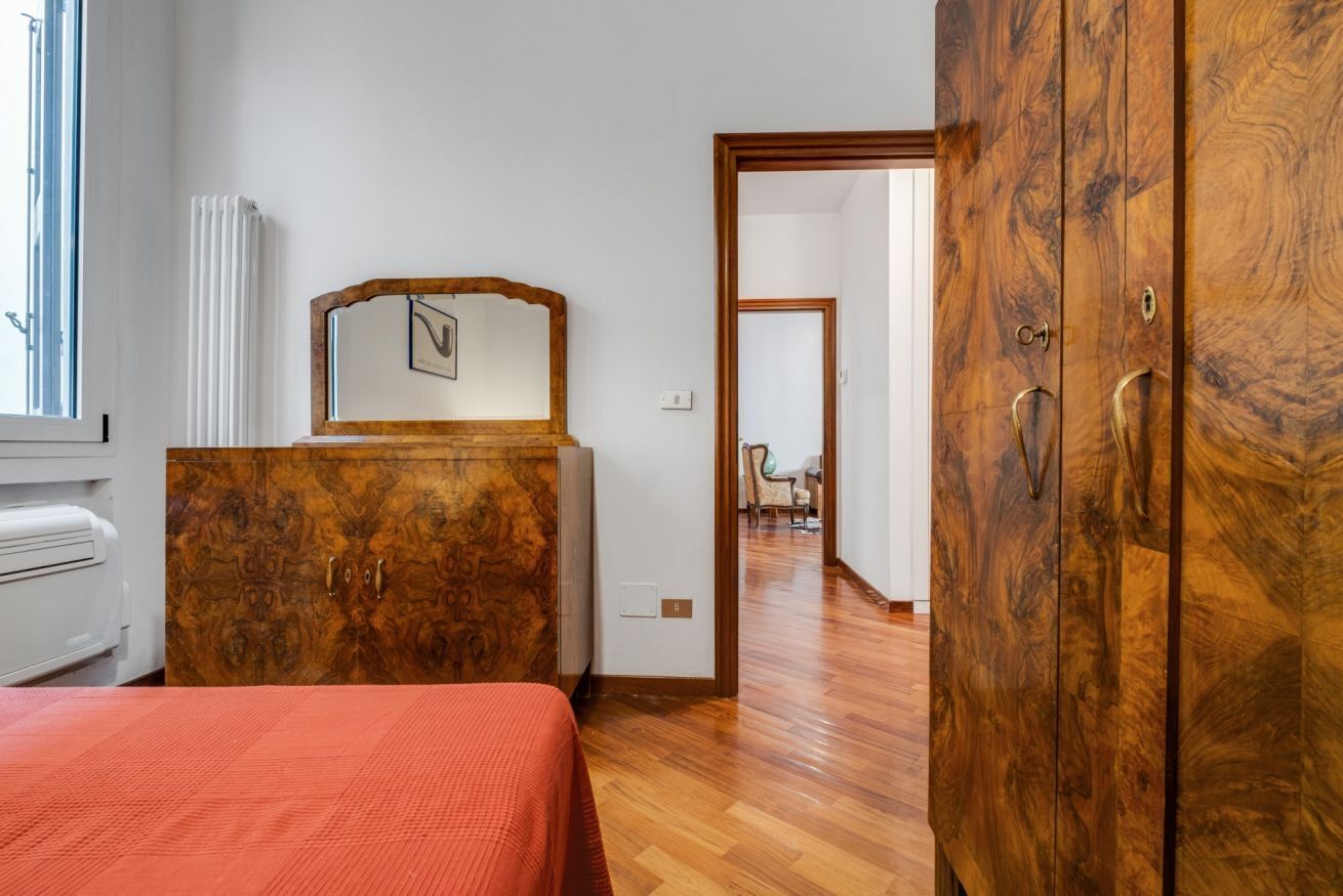 A bedroom with a wooden wardrobe, a bed with a red bedspread, a radiator, and a window.